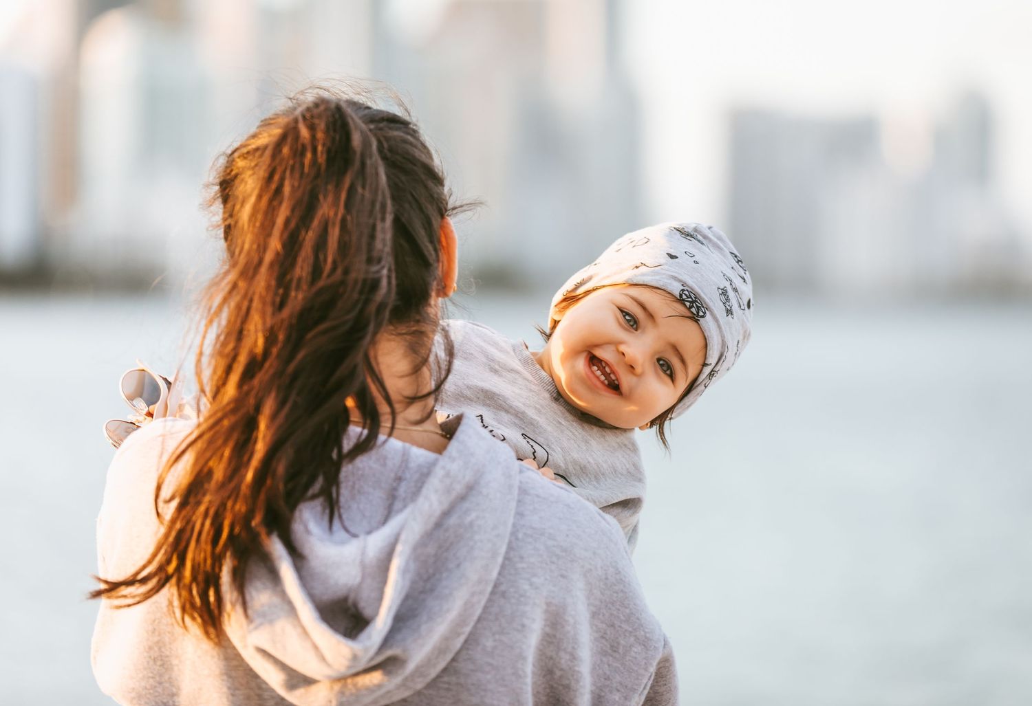 A parent in a grey hoodie holds a smiling baby wearing a beanie against a blurred cityscape background.