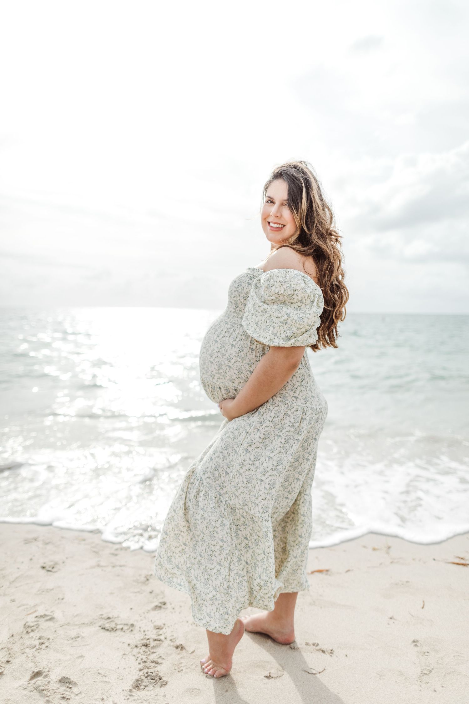 A pregnant woman in a floral dress stands barefoot on a beach with waves crashing behind her.