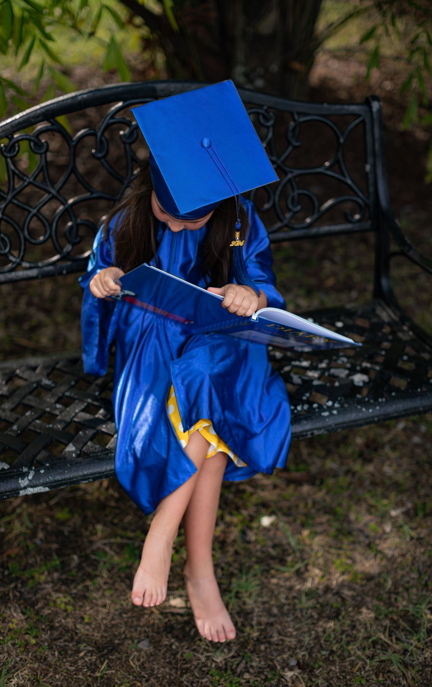 Graduate in bright blue cap and gown sits barefoot on ornate metal bench while looking at documents.