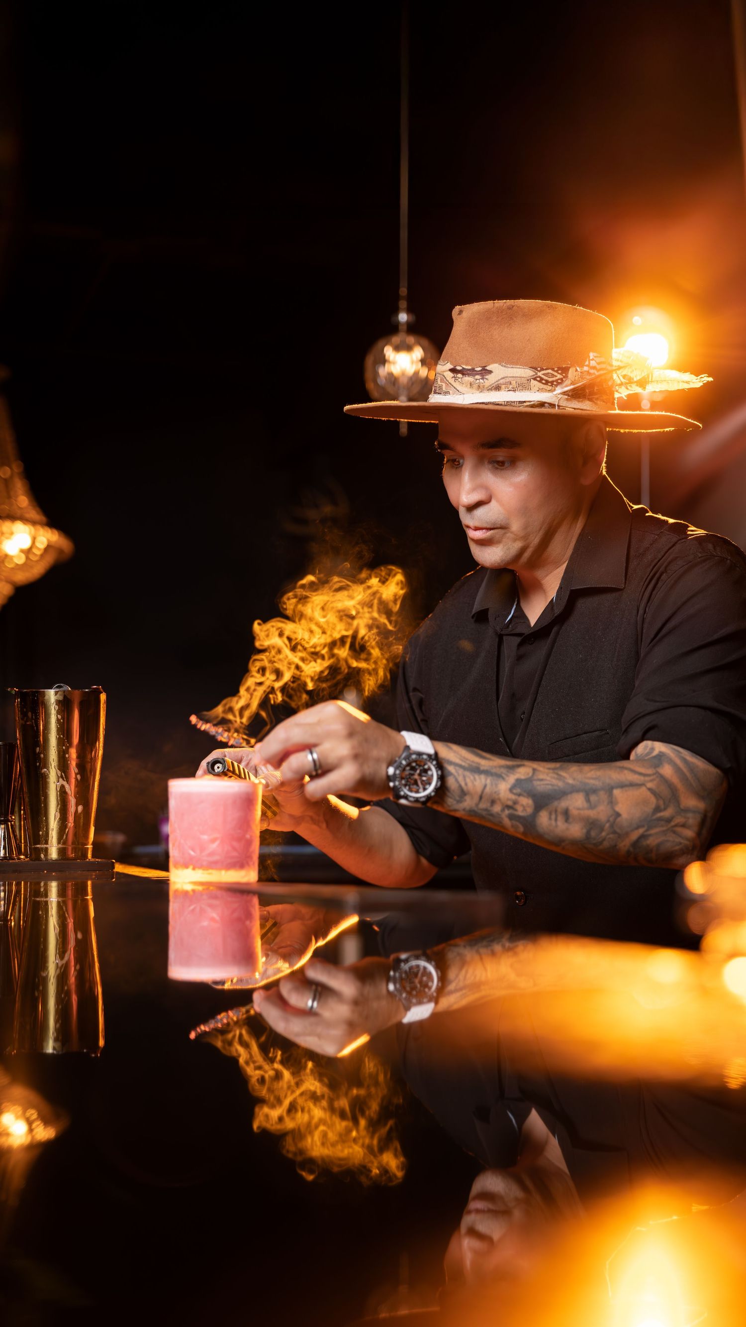 A professional bartender creates dramatic flaming cocktails with pink candles in a dark setting against orange lighting.