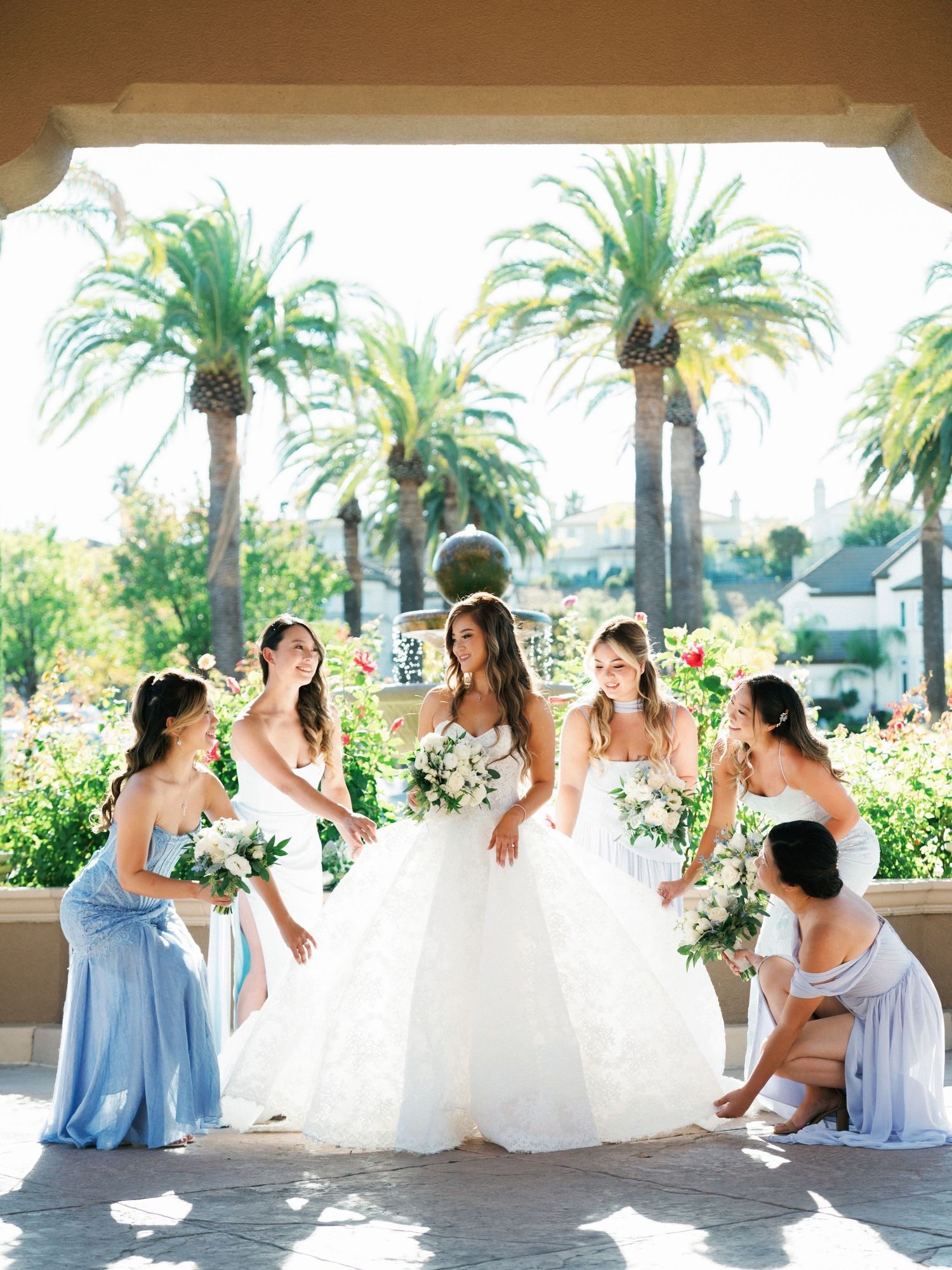 A bridal party poses outdoors with palm trees and white bouquets against a sunny tropical backdrop.