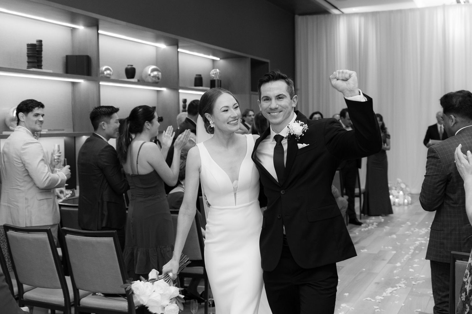 A black and white wedding reception photo showing guests mingling and dancing in an elegant venue.