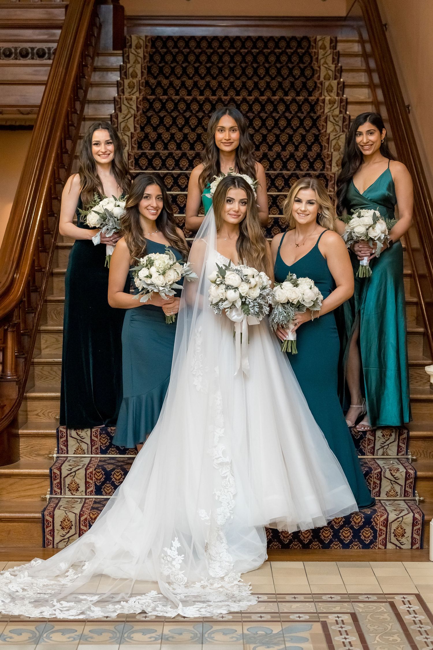 Wedding party poses in elegant teal bridesmaid dresses with white floral bouquets against ornate architectural backdrop.