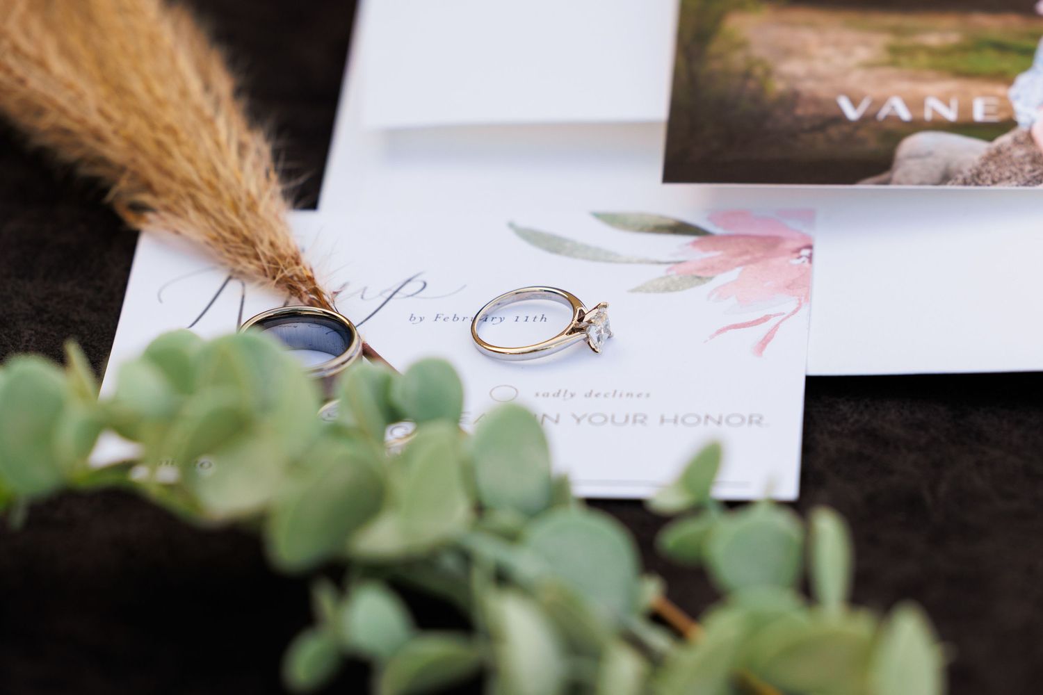 Wedding invitation details with engagement ring, dried pampas grass, and green eucalyptus leaves on dark surface.