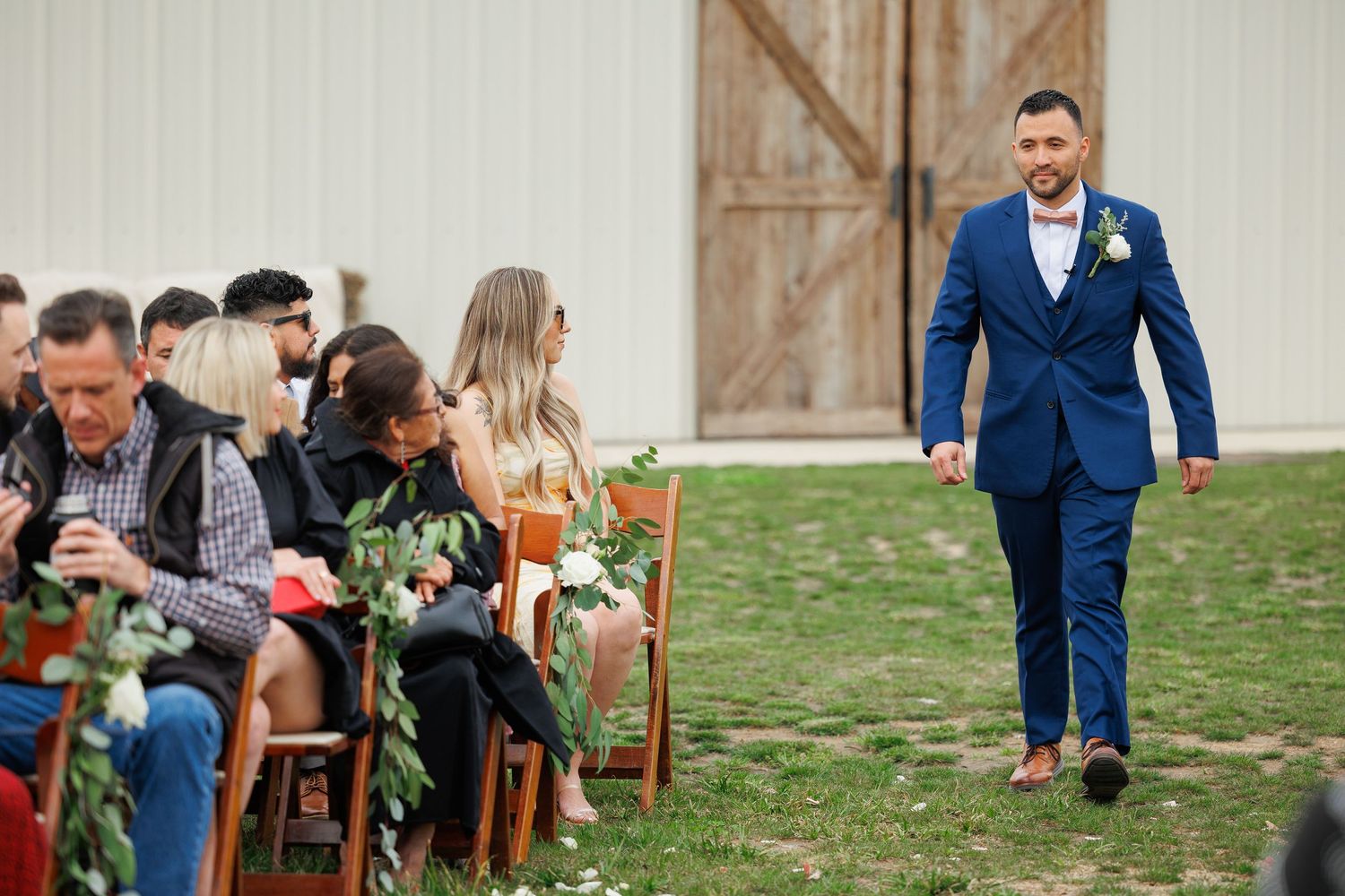 Wedding guests seated on wooden chairs at an outdoor ceremony as someone in a navy blue suit walks down the aisle.