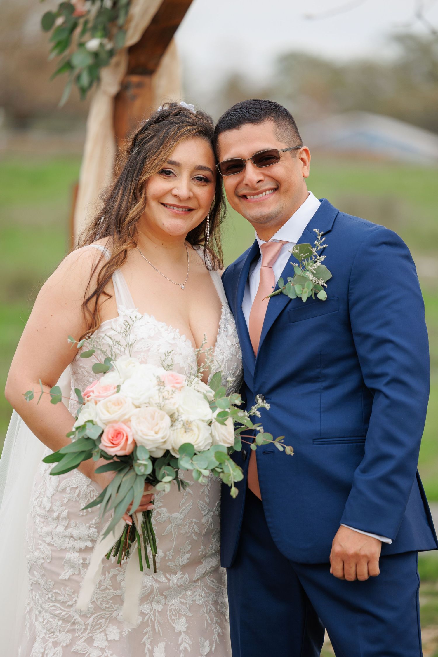 A couple in wedding attire poses together outdoors, with the bride in a lace gown and groom in a navy suit.