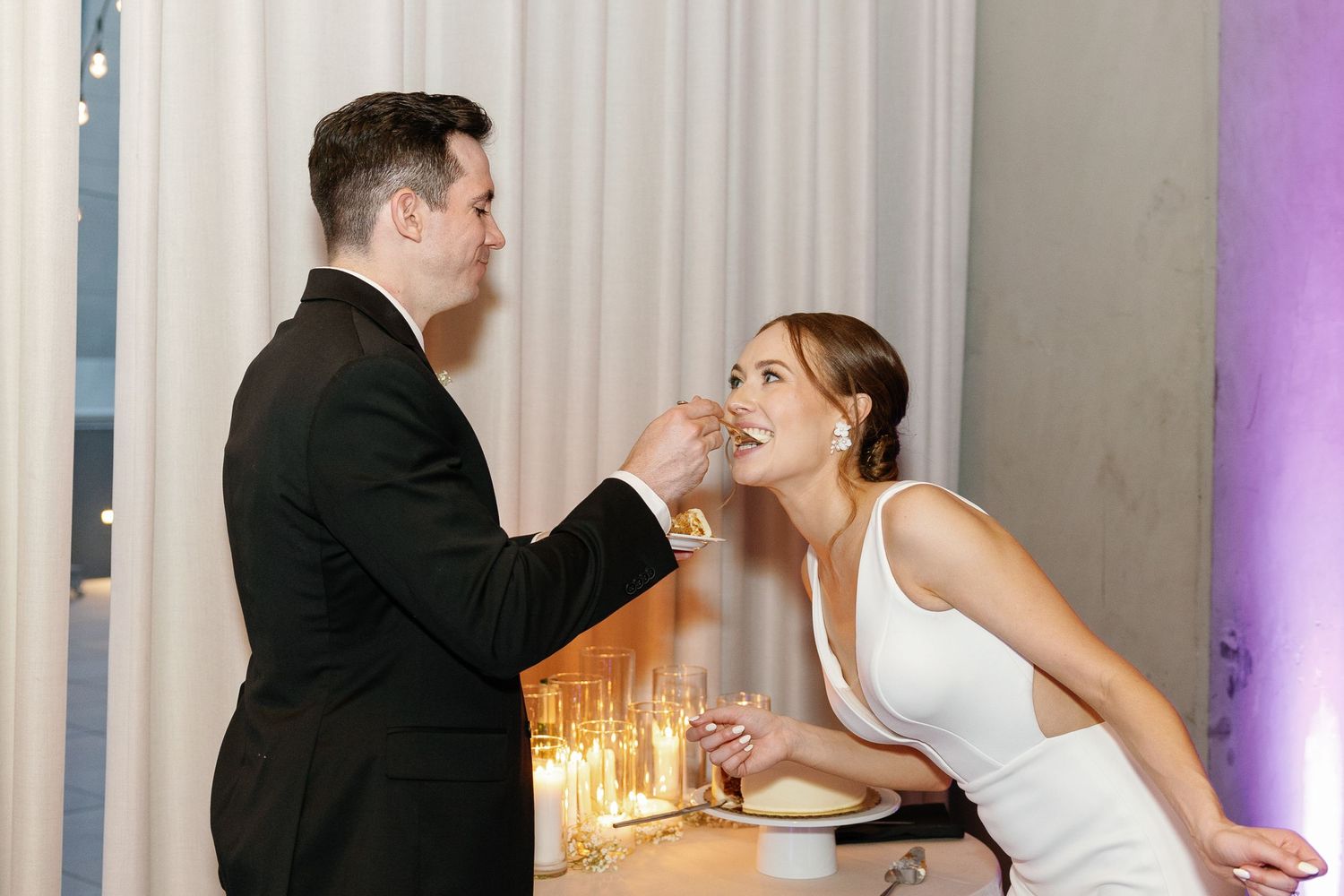 A couple shares a tender moment at their wedding reception with elegant white curtains and purple lighting in the background.