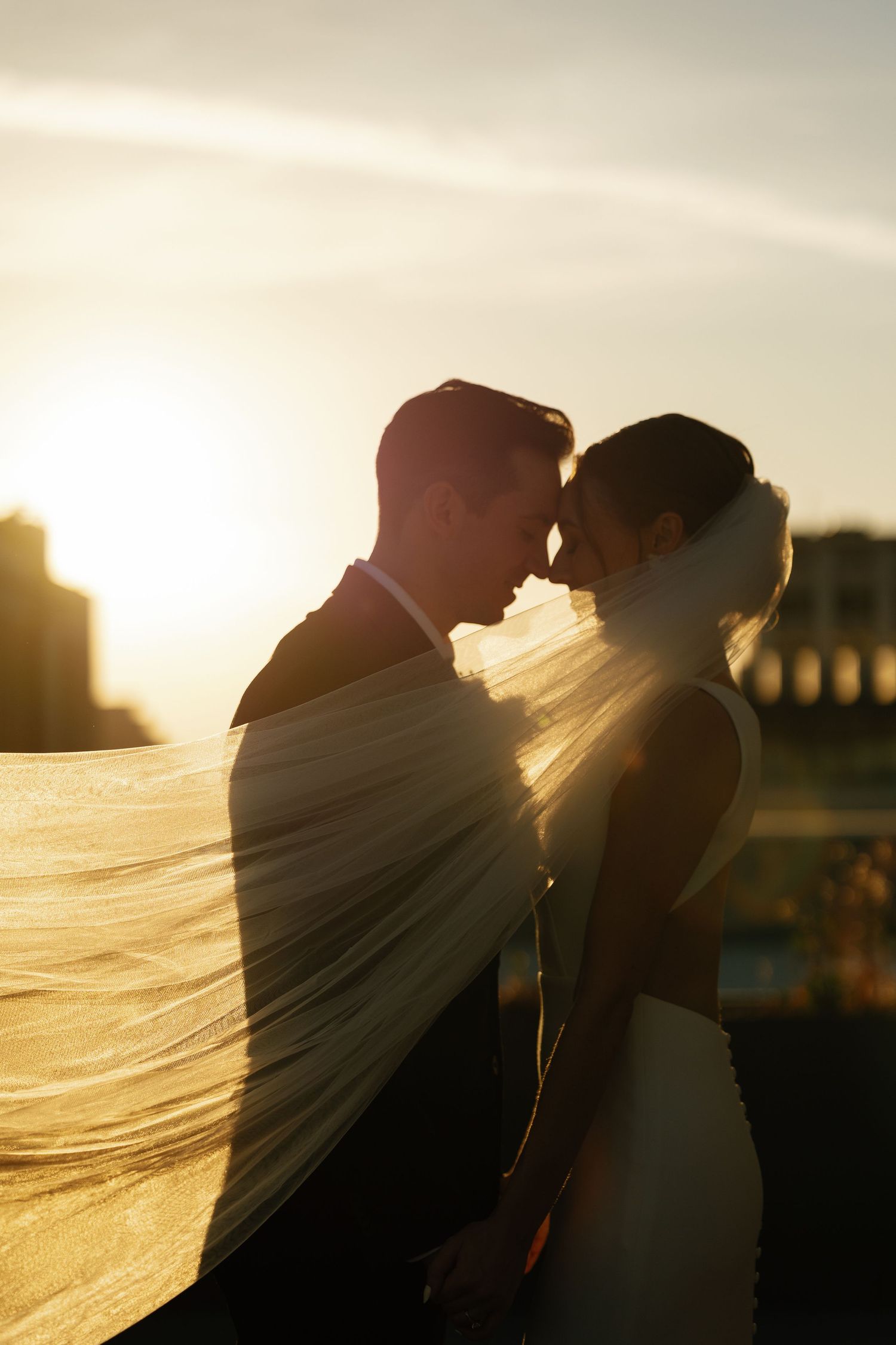 A romantic silhouette of a couple in wedding attire against a golden sunset sky with flowing veil in the breeze.