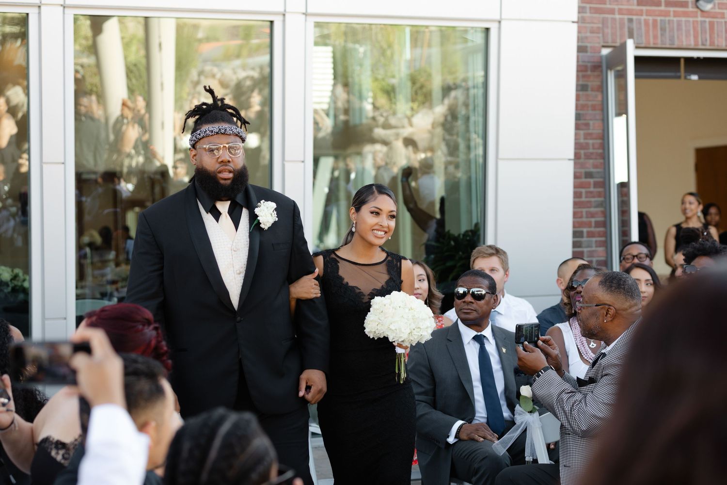 Wedding guests celebrate and take photos during an elegant outdoor ceremony at a venue with glass windows.