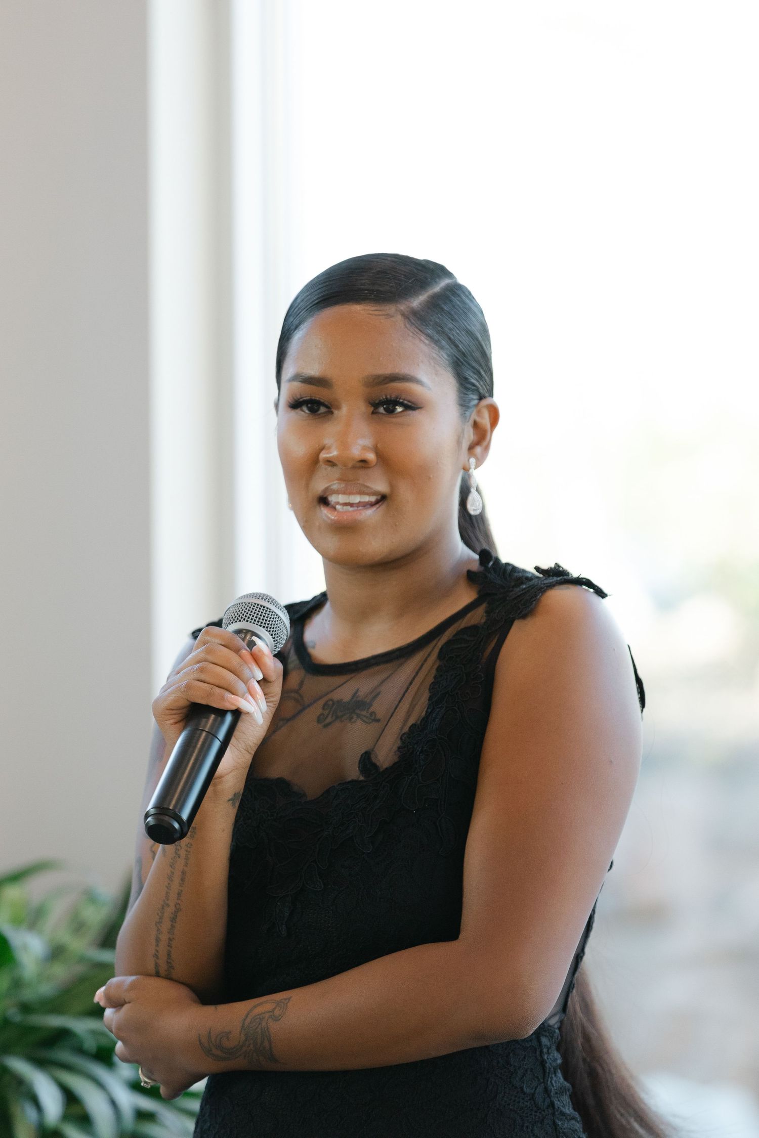 A person in a black sleeveless top smiles while holding a microphone in a bright indoor setting.