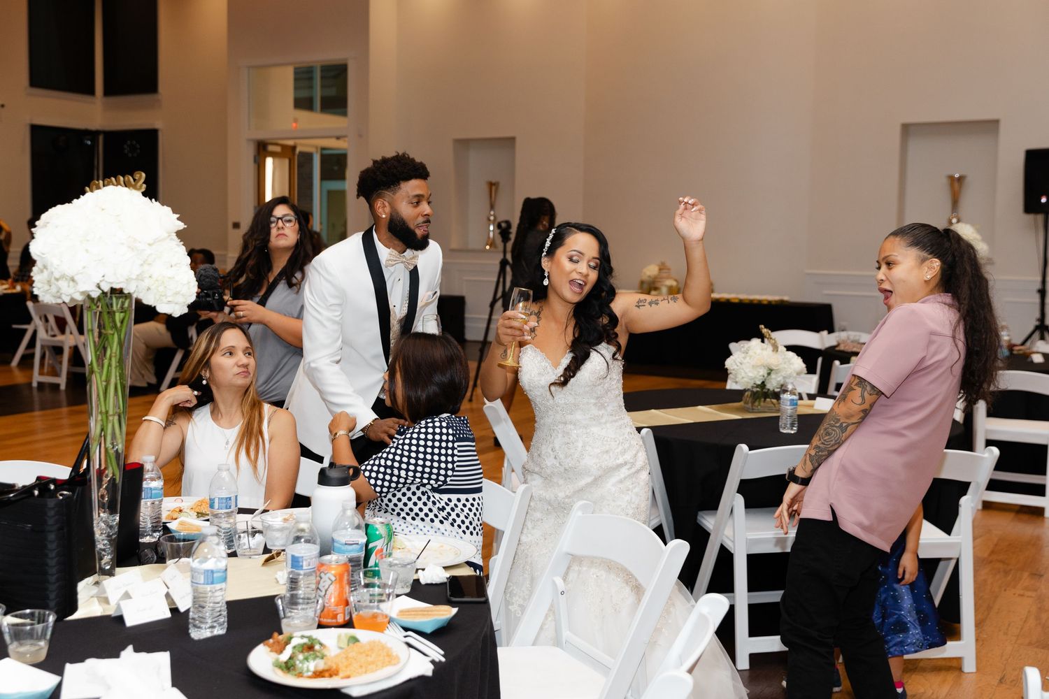 Wedding reception guests gather around a table with black linens and white decor in an elegant indoor venue.