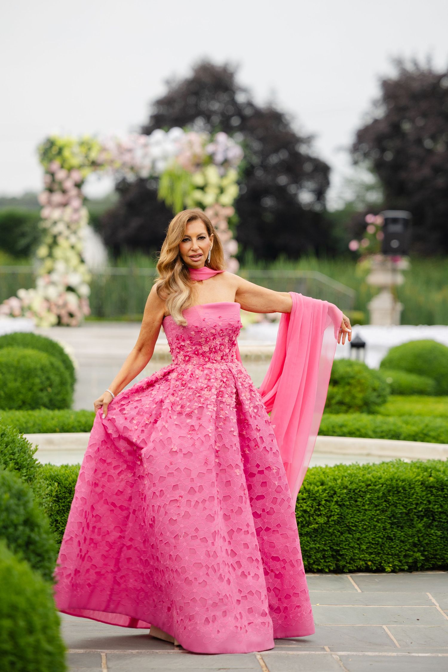 A stunning pink floral ballgown with a flowing cape is showcased in a garden setting with manicured hedges.