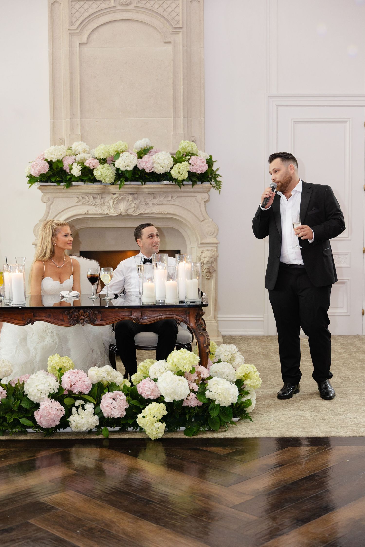 Wedding party poses for creative photo series against elegant white mantle decorated with pink and white flowers and candles.