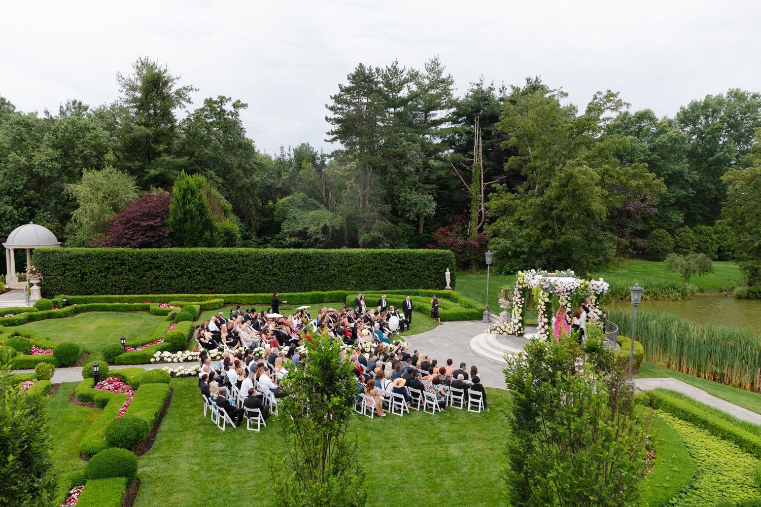 Aerial view of an outdoor garden wedding ceremony with guests seated in white chairs surrounded by manicured hedges and trees.
