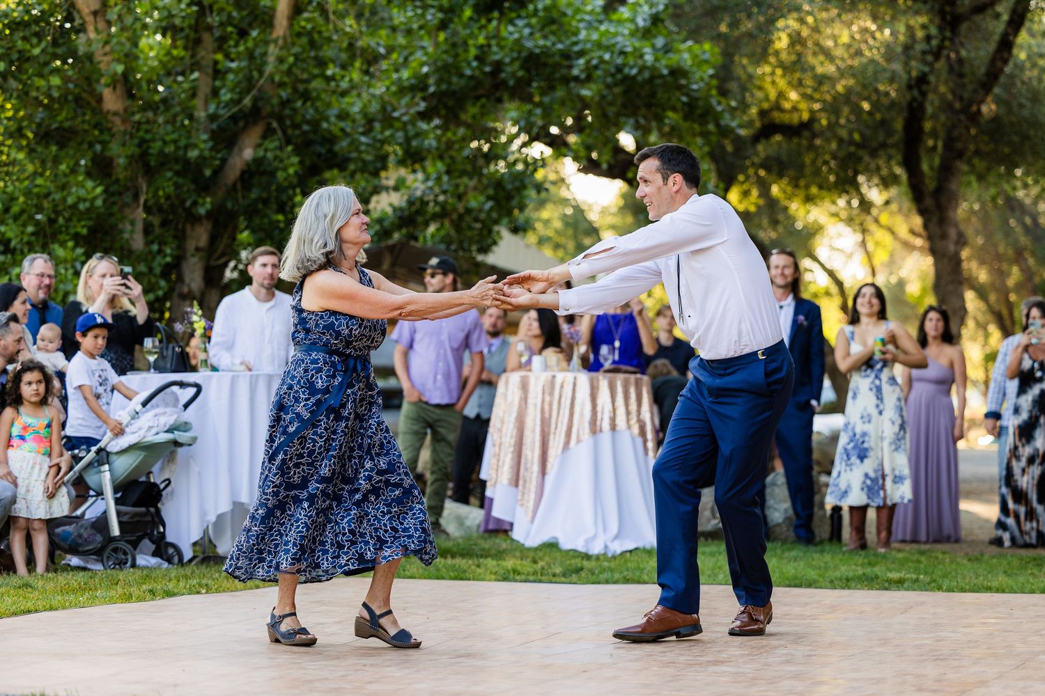 Elderly couple dancing together at an outdoor garden party with guests in formal attire watching in the background.