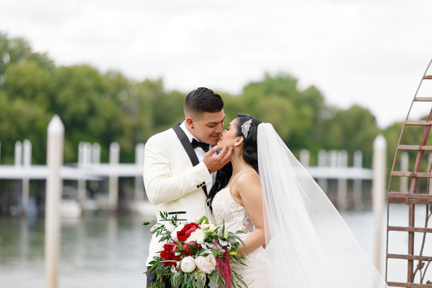 Newlyweds share a romantic kiss on a wooden dock overlooking a scenic waterfront with boats in the background.