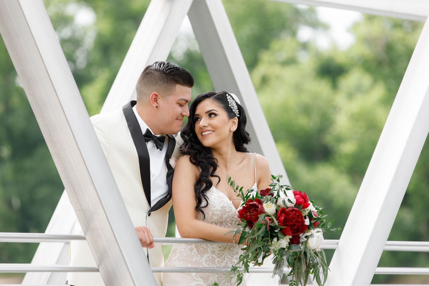 Newlywed couple shares intimate moment on white bridge with bride holding vibrant red bouquet.