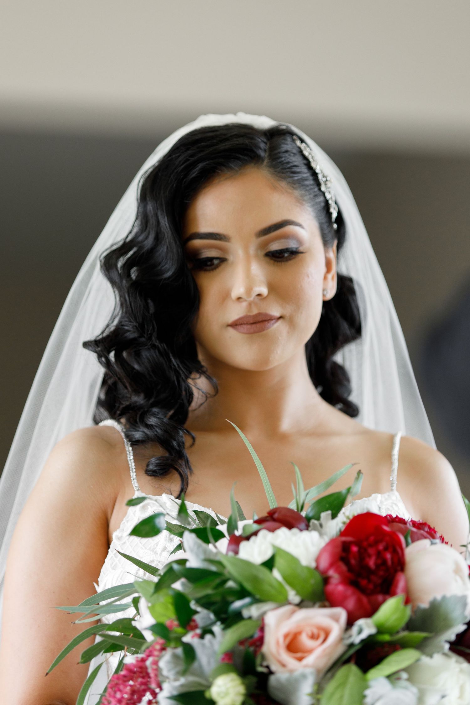 A bride in a white veil holds a vibrant red and pink rose bouquet while smiling softly in a bridal portrait.