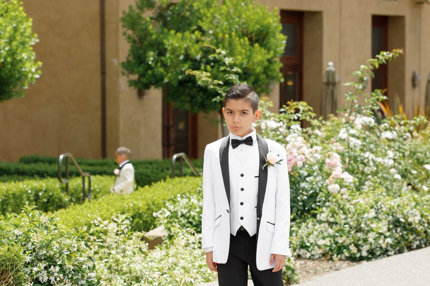 A person in a white tuxedo jacket stands in a garden with blooming roses and adobe buildings in the background.