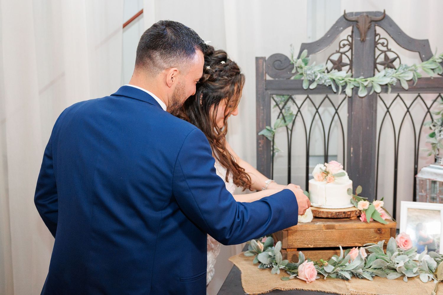 A couple in blue attire signs documents together at a wooden table decorated with greenery and floral arrangements.