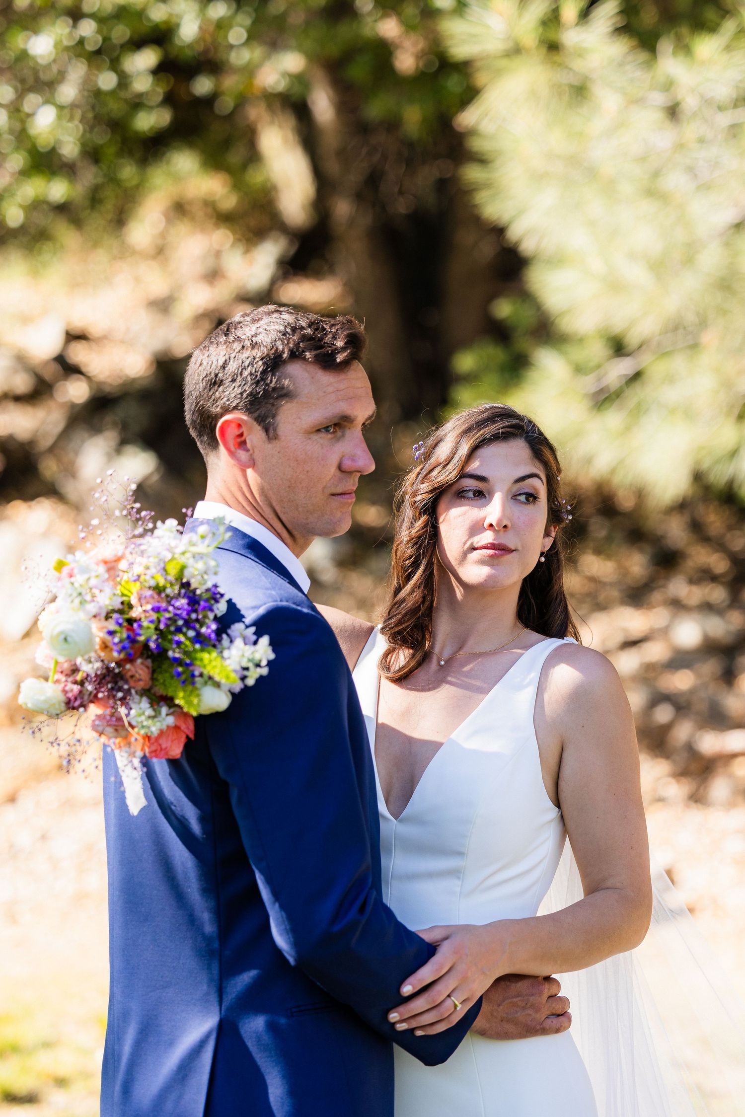 A couple in wedding attire embraces outdoors, with the bride in white and groom in navy blue holding a colorful bouquet.