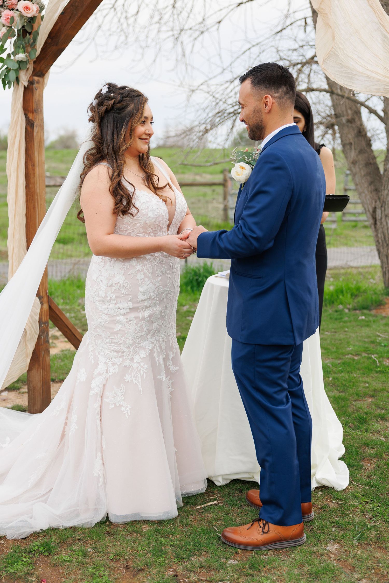 A couple in wedding attire shares a playful first look moment outdoors under a rustic wooden arch.