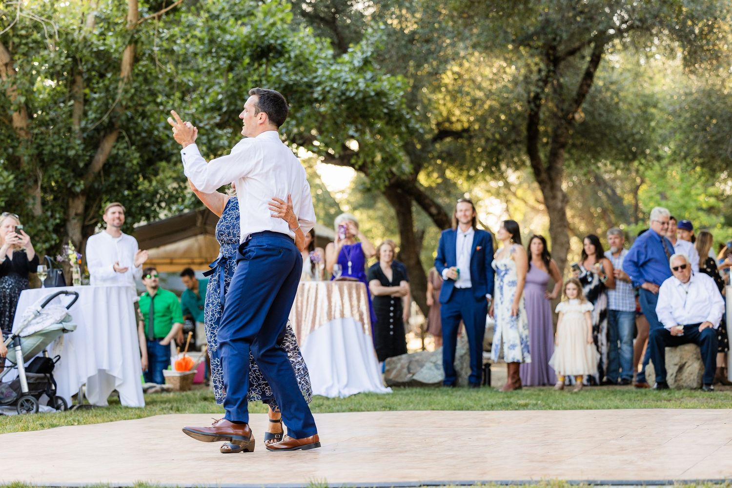 Wedding guests gather outdoors under shady trees for a summer celebration in a natural setting.