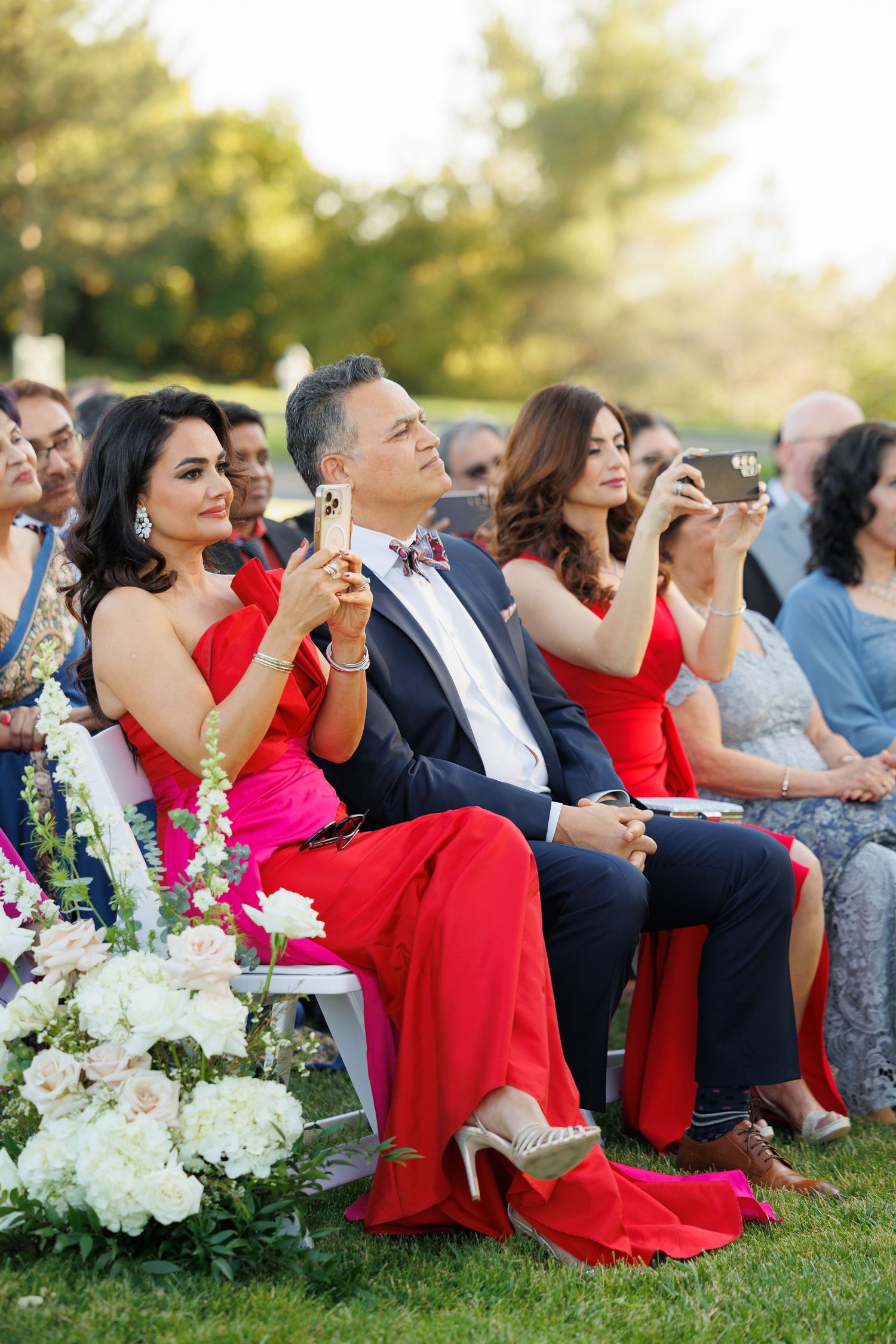 Wedding guests in red dresses and formal attire sit on grass watching an outdoor ceremony.