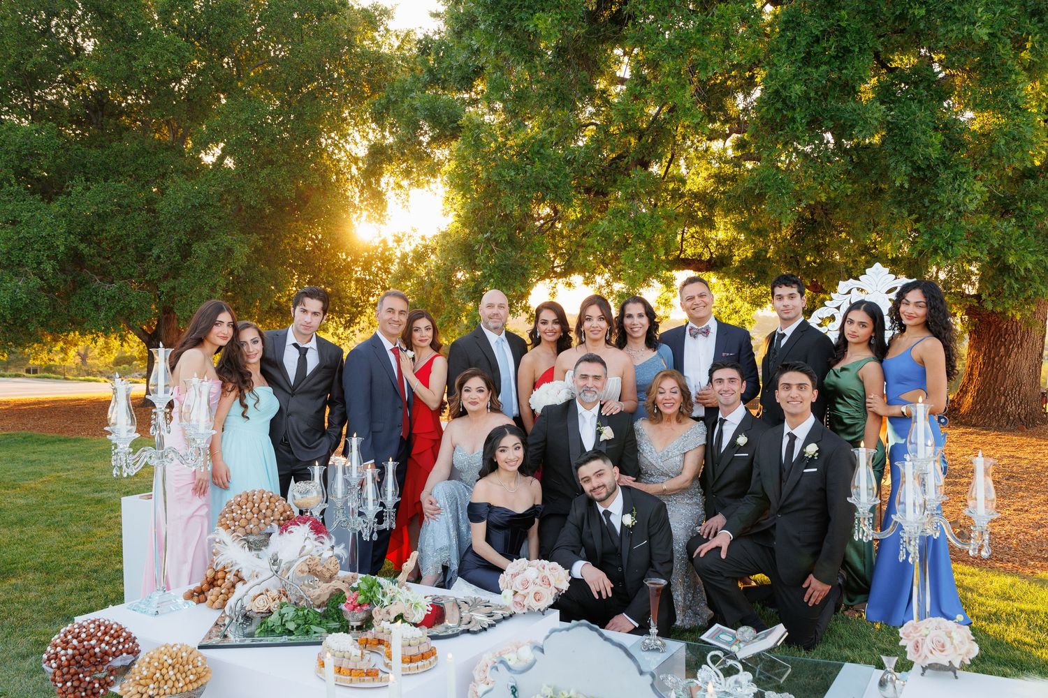 Wedding party gathers for outdoor photos during golden hour sunset in a lush garden setting.