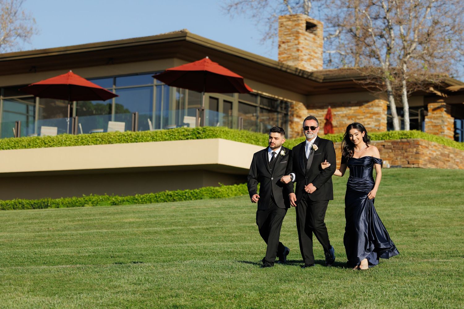 Three people in formal attire walk across a lush green lawn in front of a modern building with large windows.