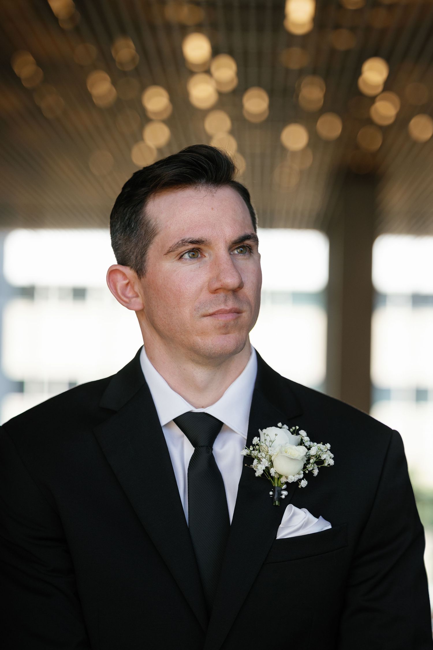 A groom in a black suit and white tie looks thoughtfully to the side with a white boutonniere.