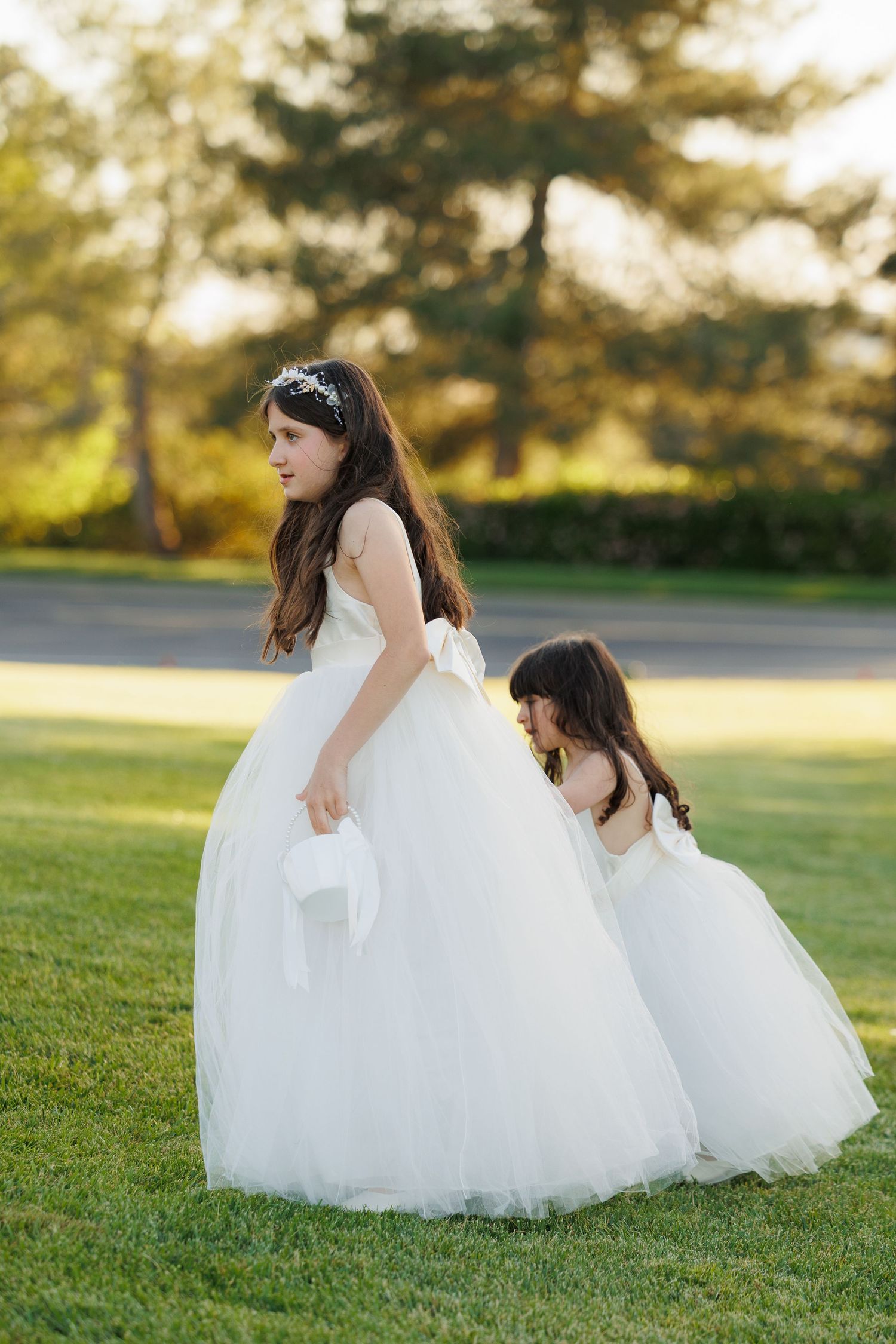Two figures in flowing white dresses sit together on green grass during golden hour.