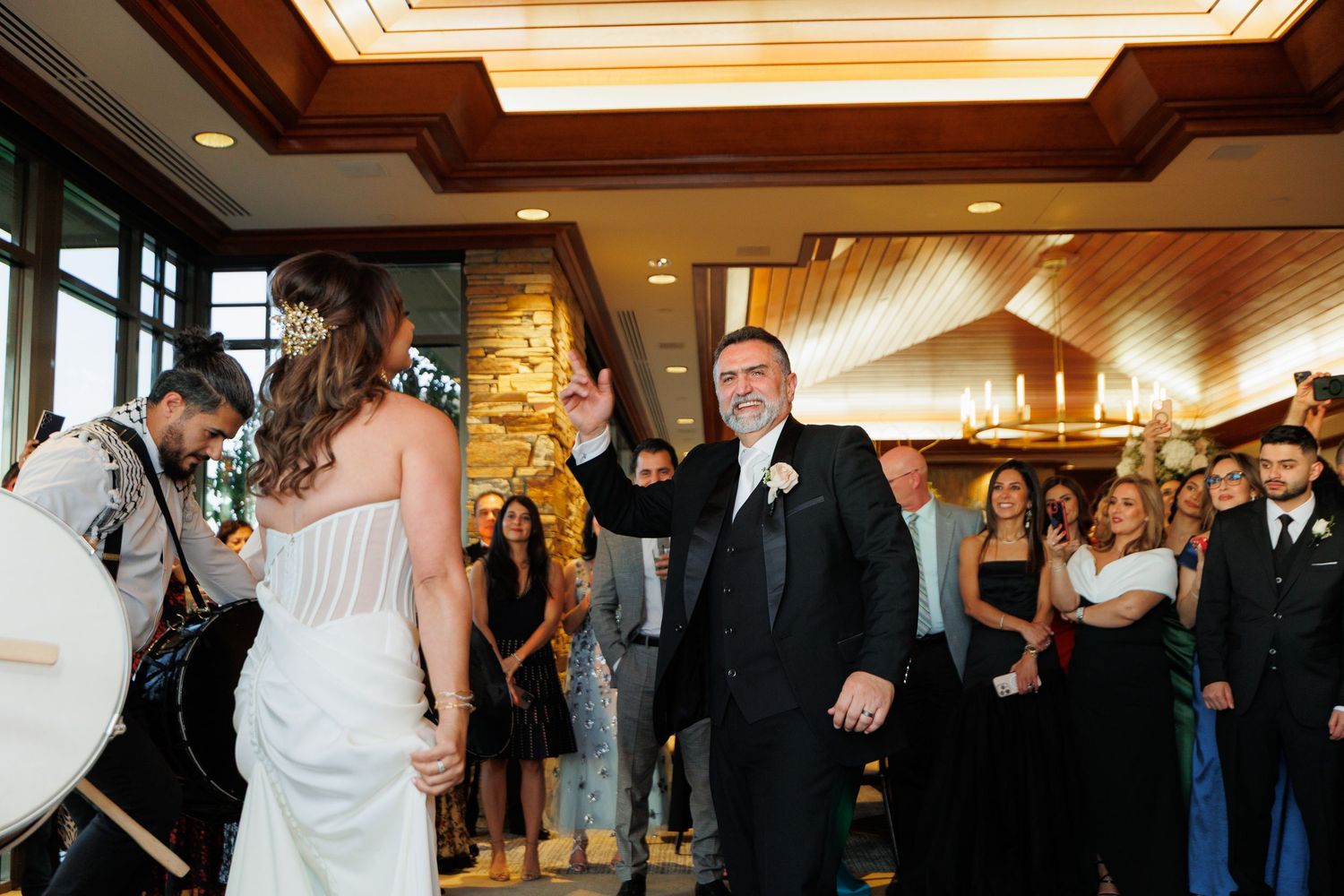 A group of wedding guests dance and celebrate together in an elegant ballroom with wooden ceiling details.