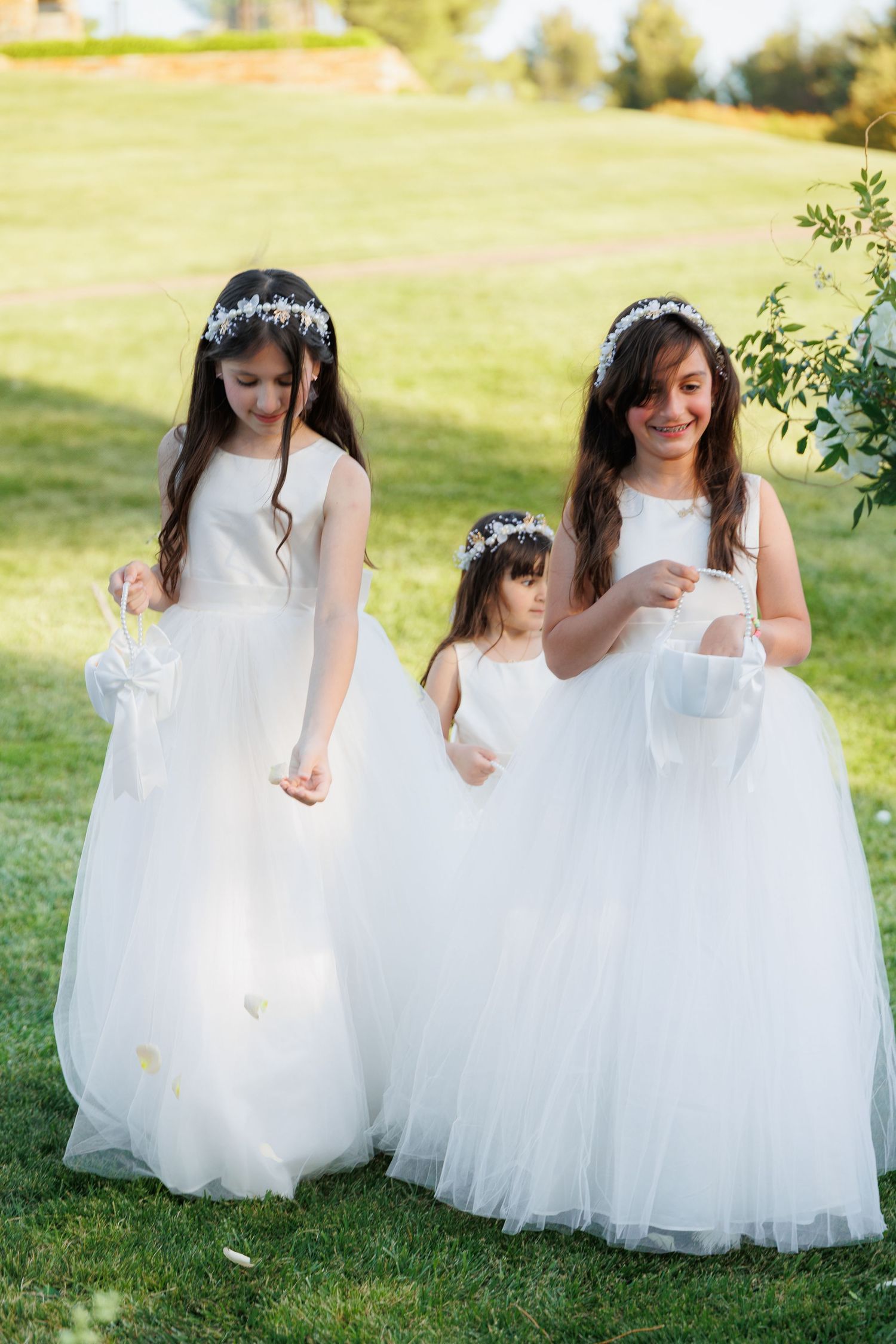Two flower girls in white dresses twirl and play together on a sunny green lawn during a wedding celebration.