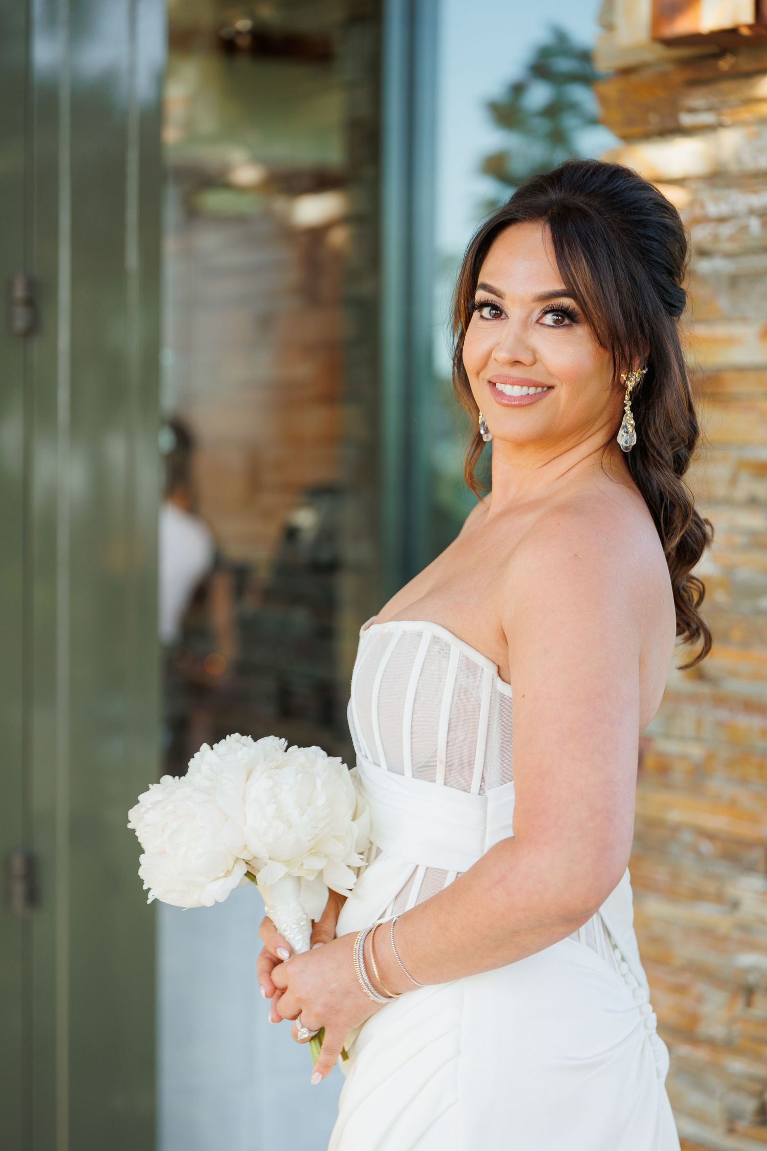 Beautiful bride in white strapless wedding dress holds white peony bouquet while smiling against brick wall backdrop.