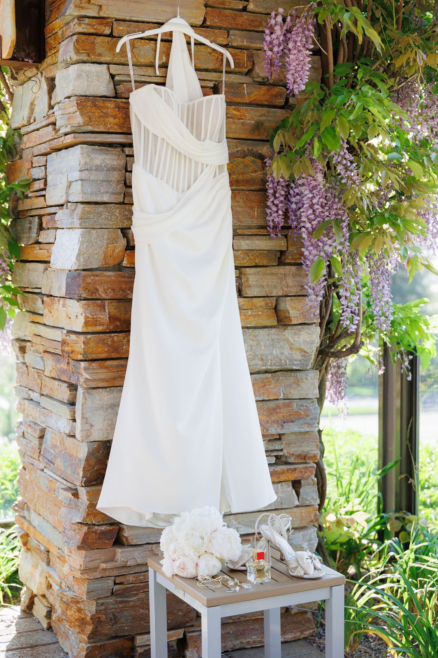 White wedding dress displayed on hangers against rustic stone wall with purple wisteria flowers hanging above.