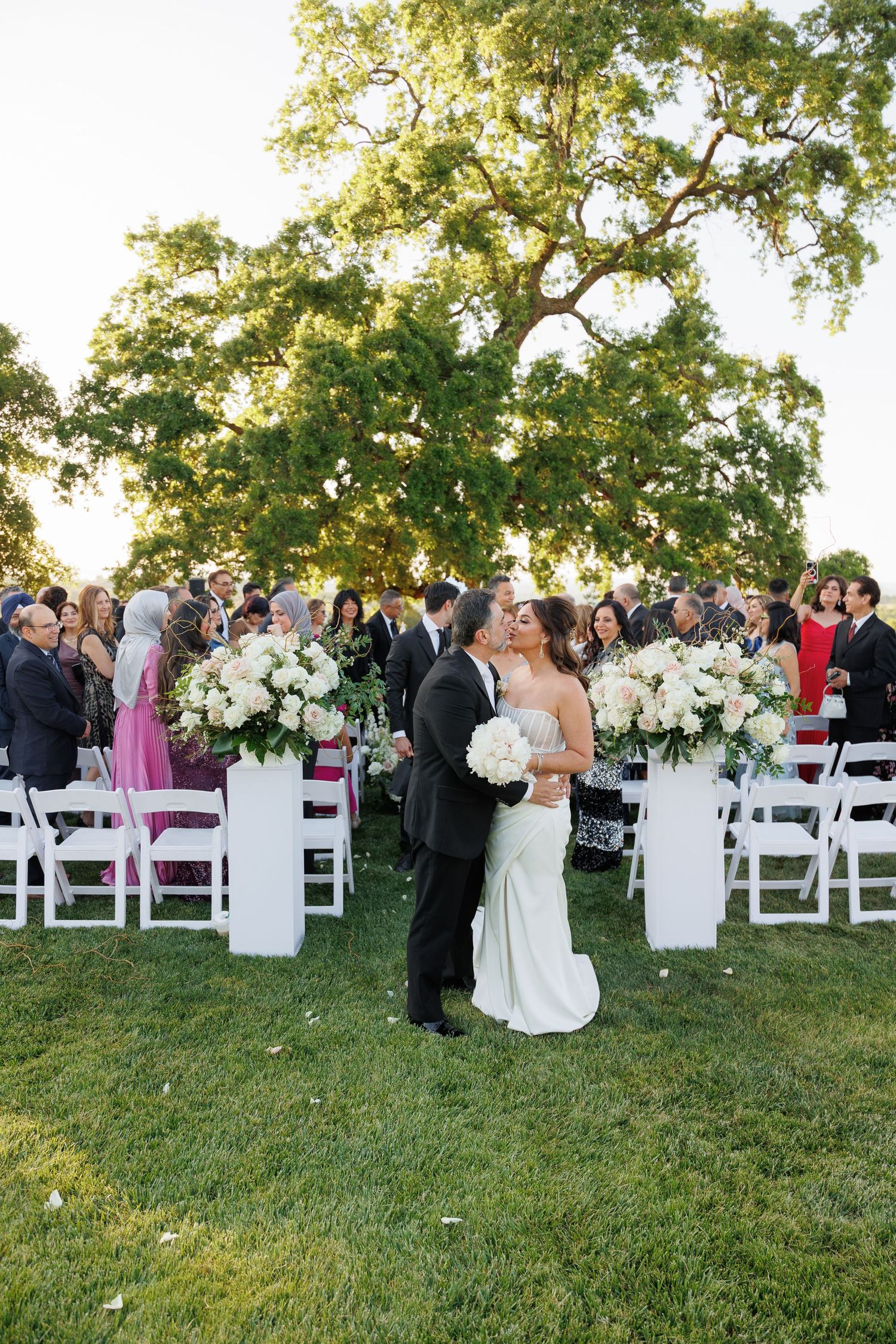 Wedding ceremony guests gather on a grassy lawn with white chairs and floral arrangements at an outdoor venue.