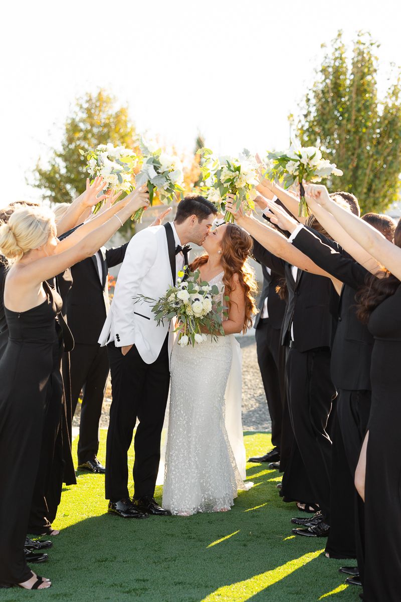 Wedding party members in black attire perform synchronized dab dance pose on sunny lawn during celebration.