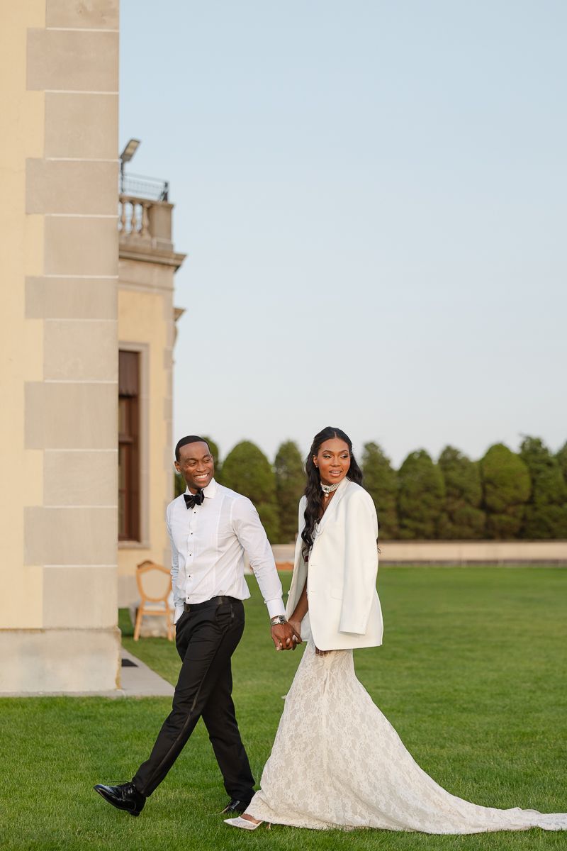 A couple in wedding attire walks playfully across a grand lawn with stone architecture in the background.
