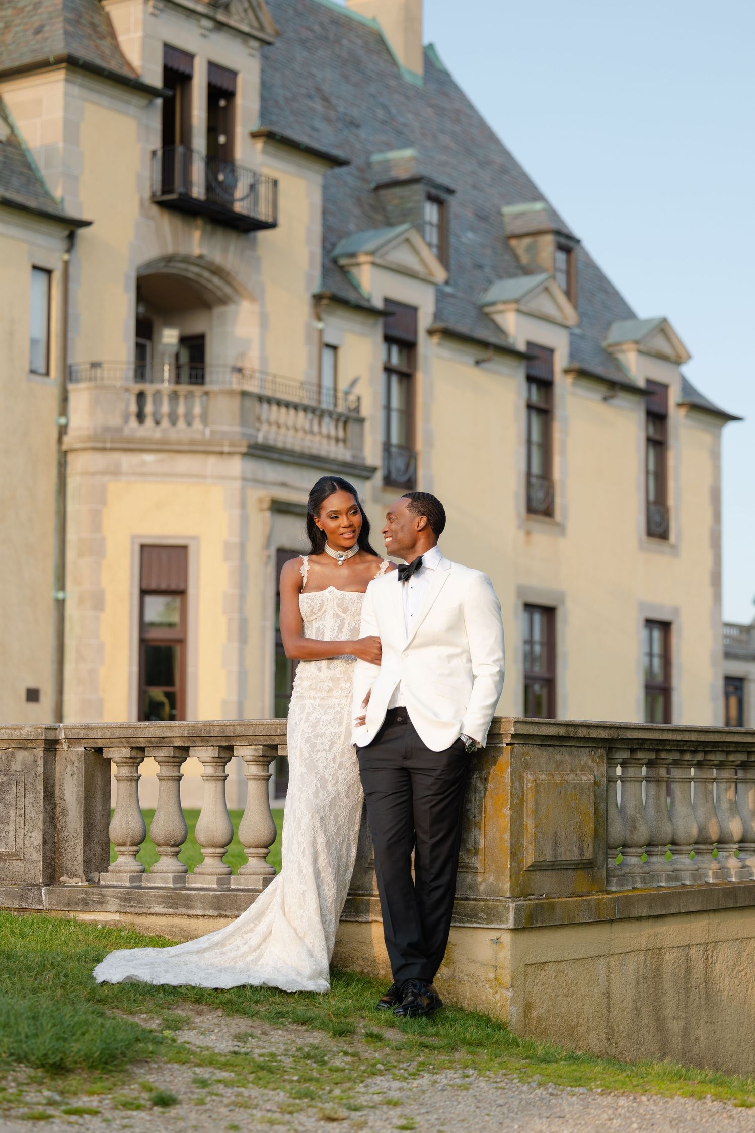 A wedding couple poses elegantly in front of a grand stone mansion with classic architecture.