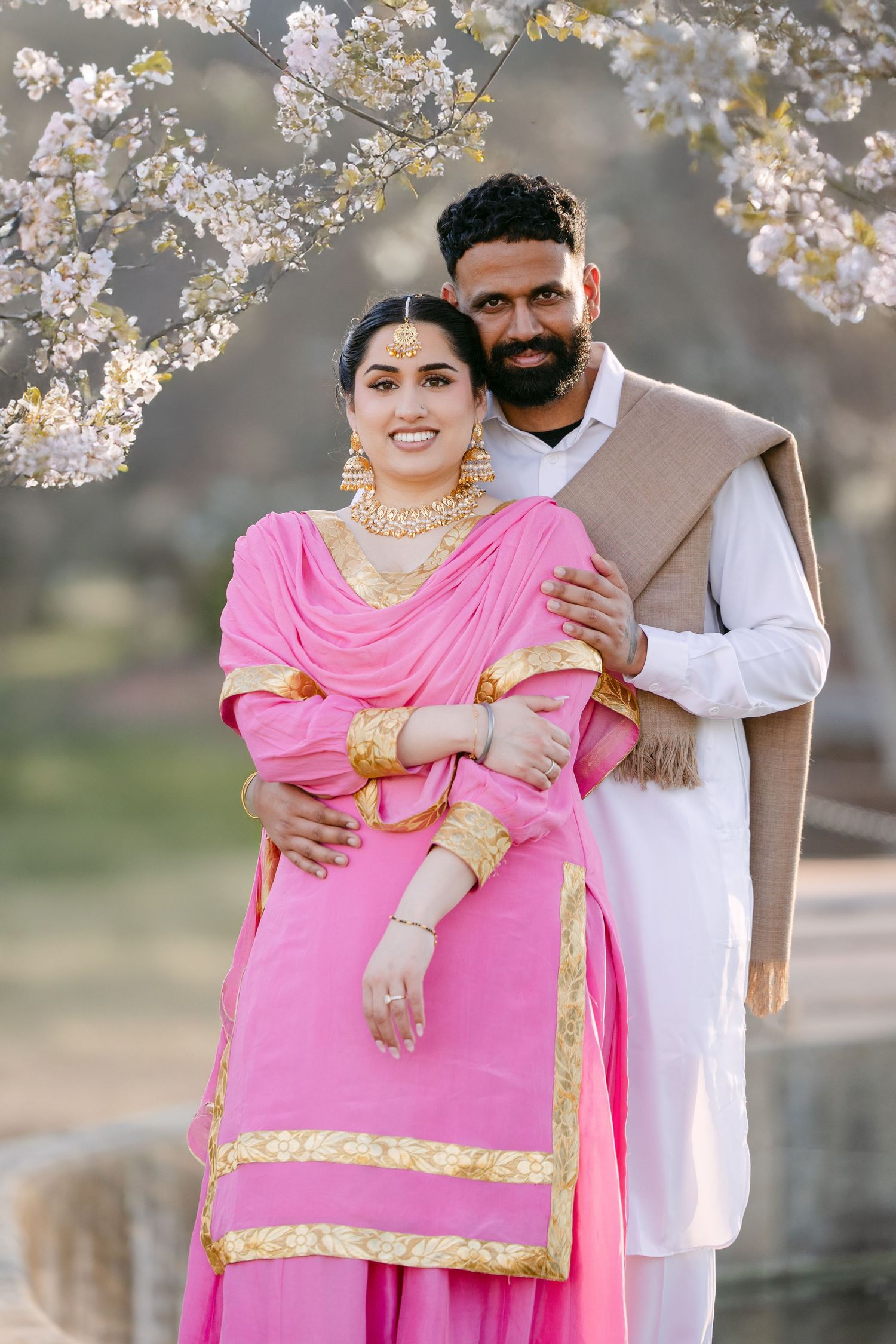 A couple in traditional Indian attire poses together under blooming cherry blossom trees during a spring photoshoot.