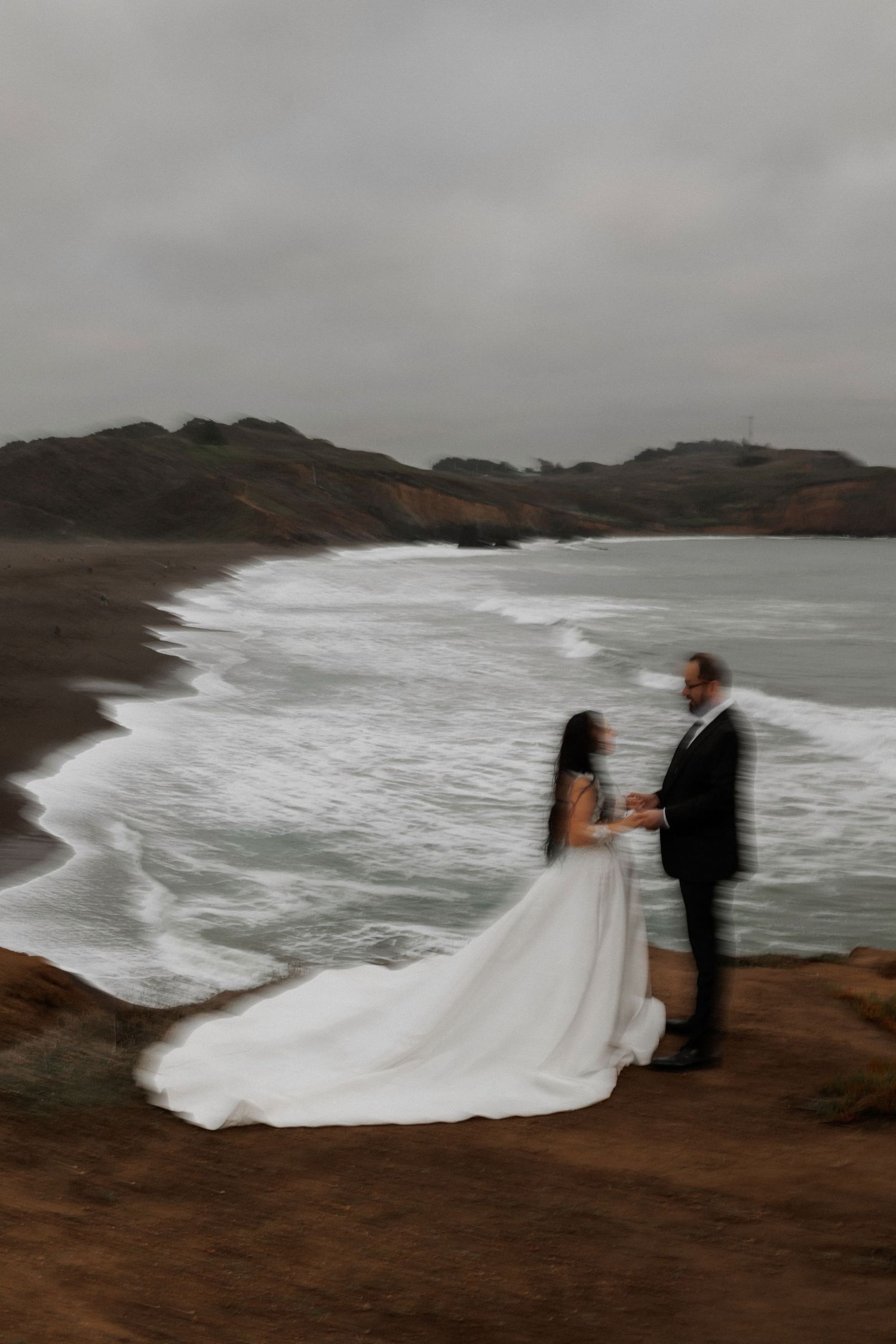 A couple in wedding attire stands on a cliff overlooking dramatic ocean waves on a cloudy day.