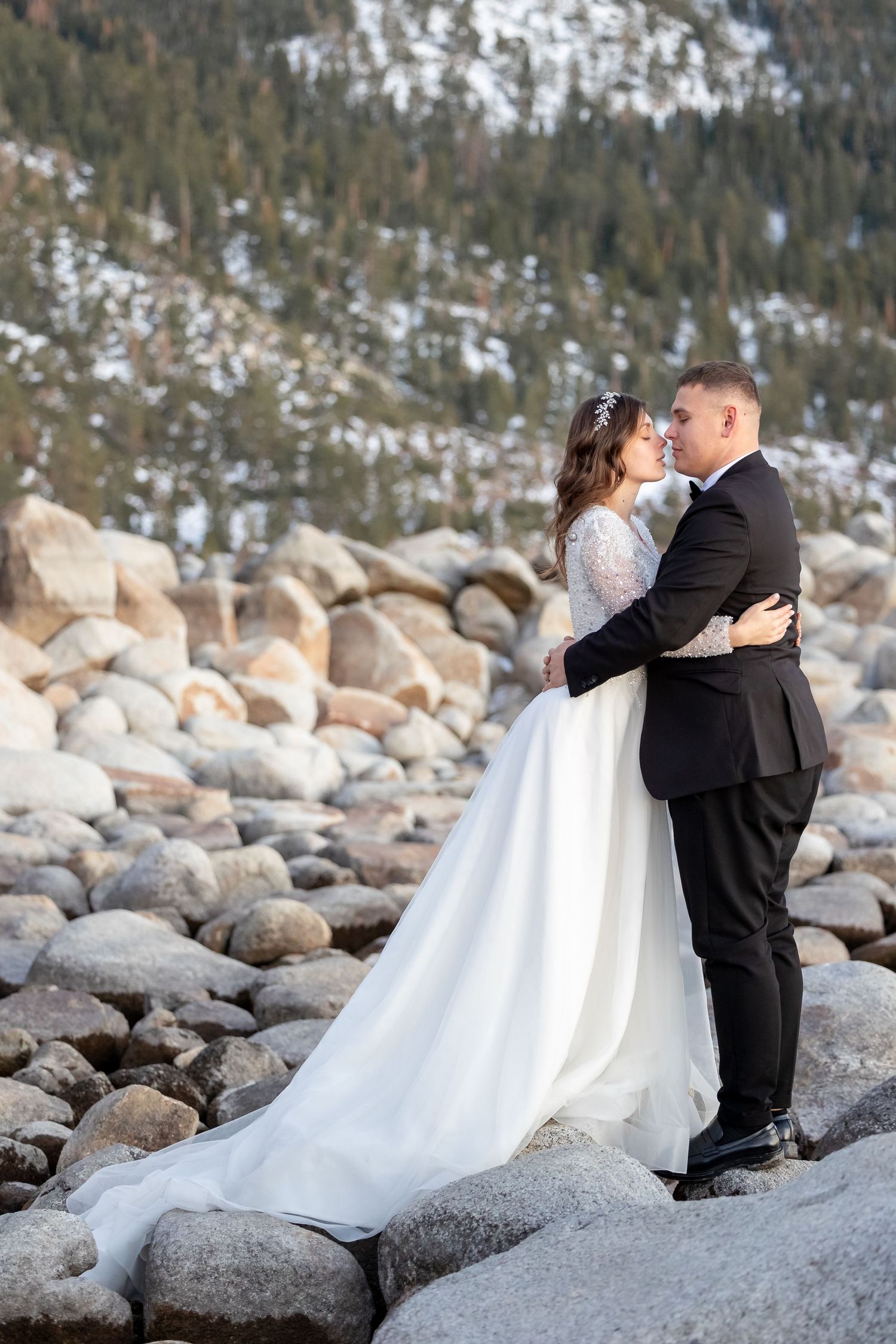Winter wedding photography session on snowy rocky shoreline with mountains in background.