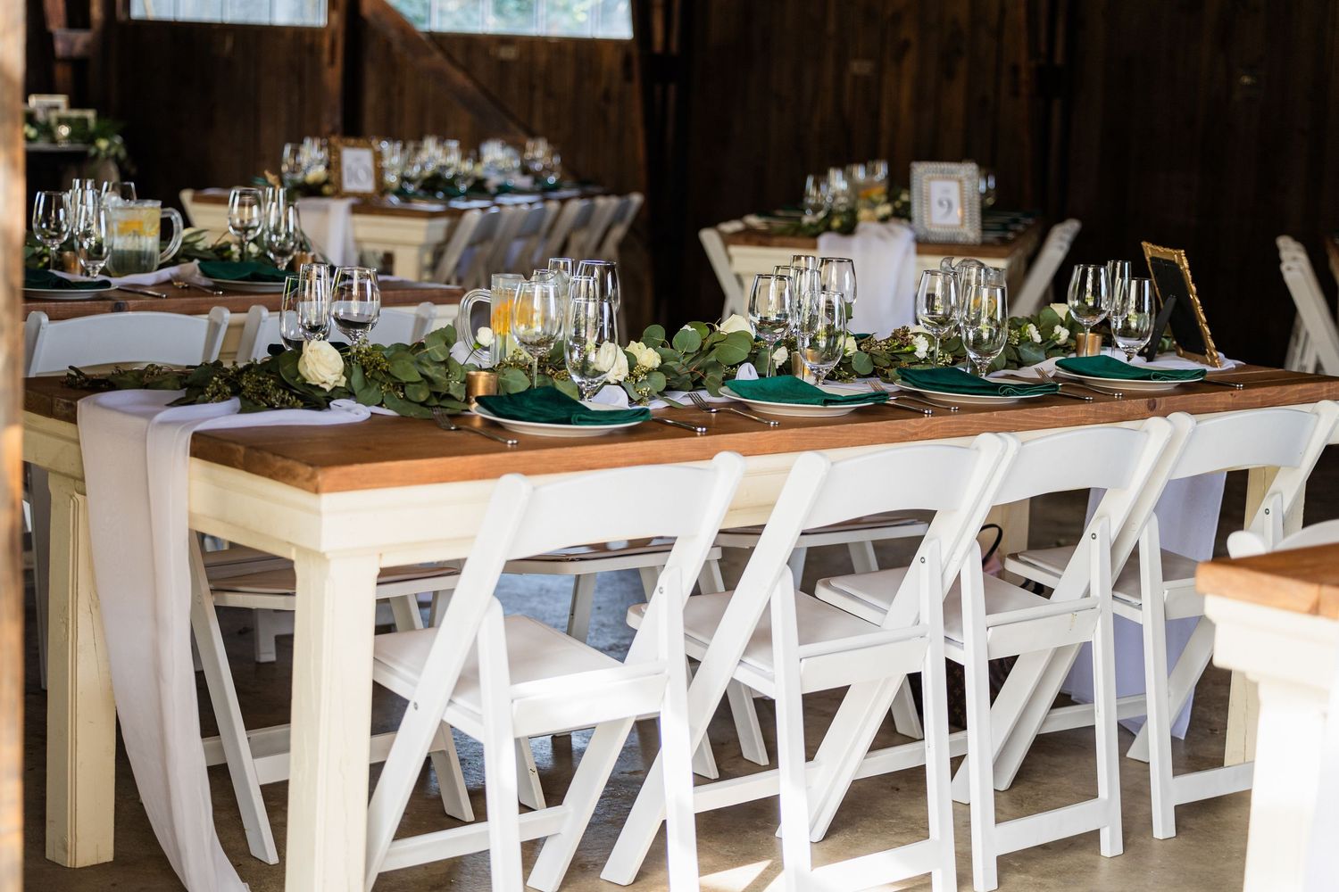 Wedding reception hall with white chairs and long tables decorated with greenery and elegant place settings.