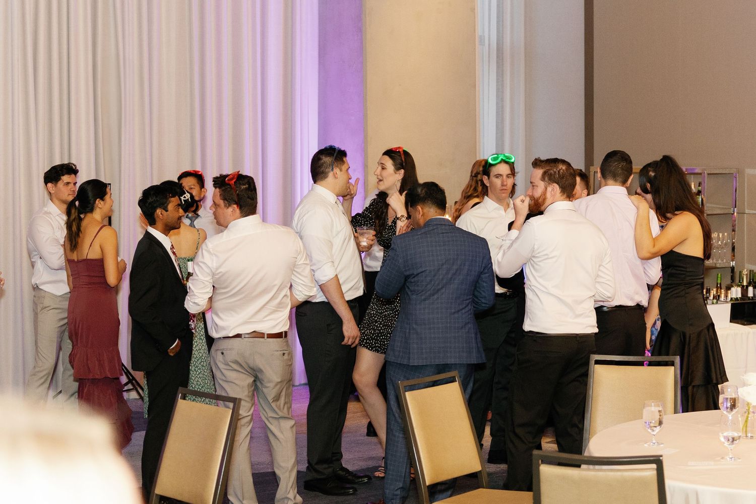 A group of guests mingle and socialize at an elegant event with purple uplighting and cream-colored chairs.