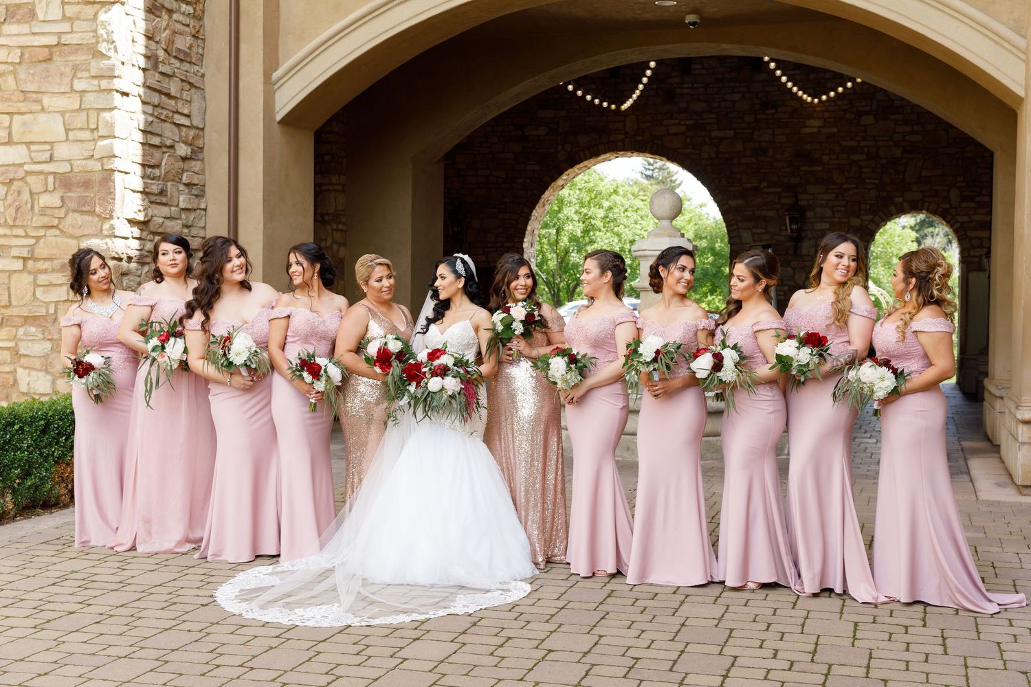Wedding party in pink bridesmaid dresses pose together under stone archways during an outdoor celebration.