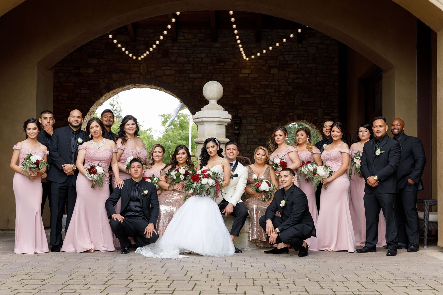 A wedding party poses together under an arched stone entryway wearing pink bridesmaid dresses and formal attire.