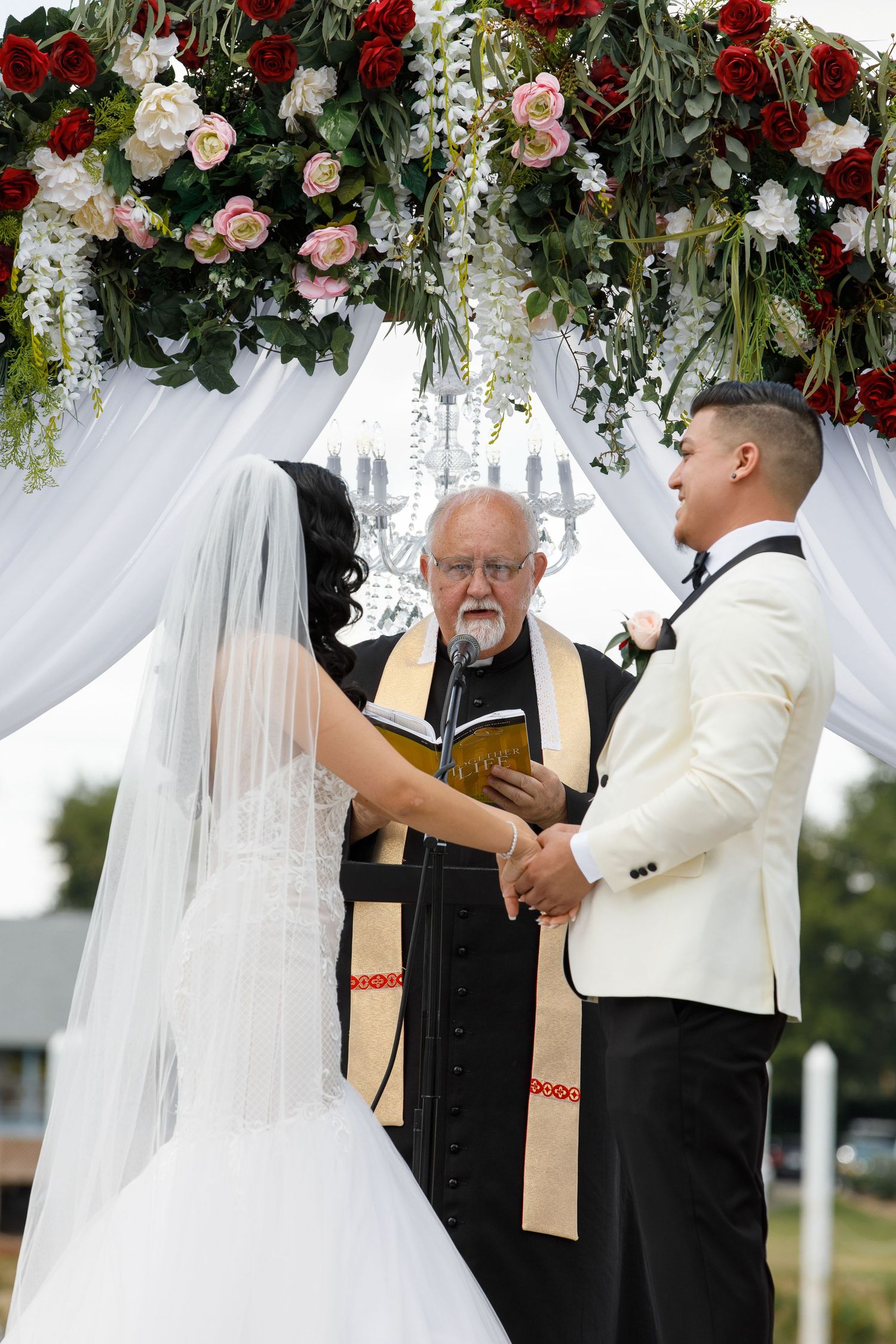 Wedding ceremony under floral arch with red and pink roses and white wisteria with officiant conducting service.