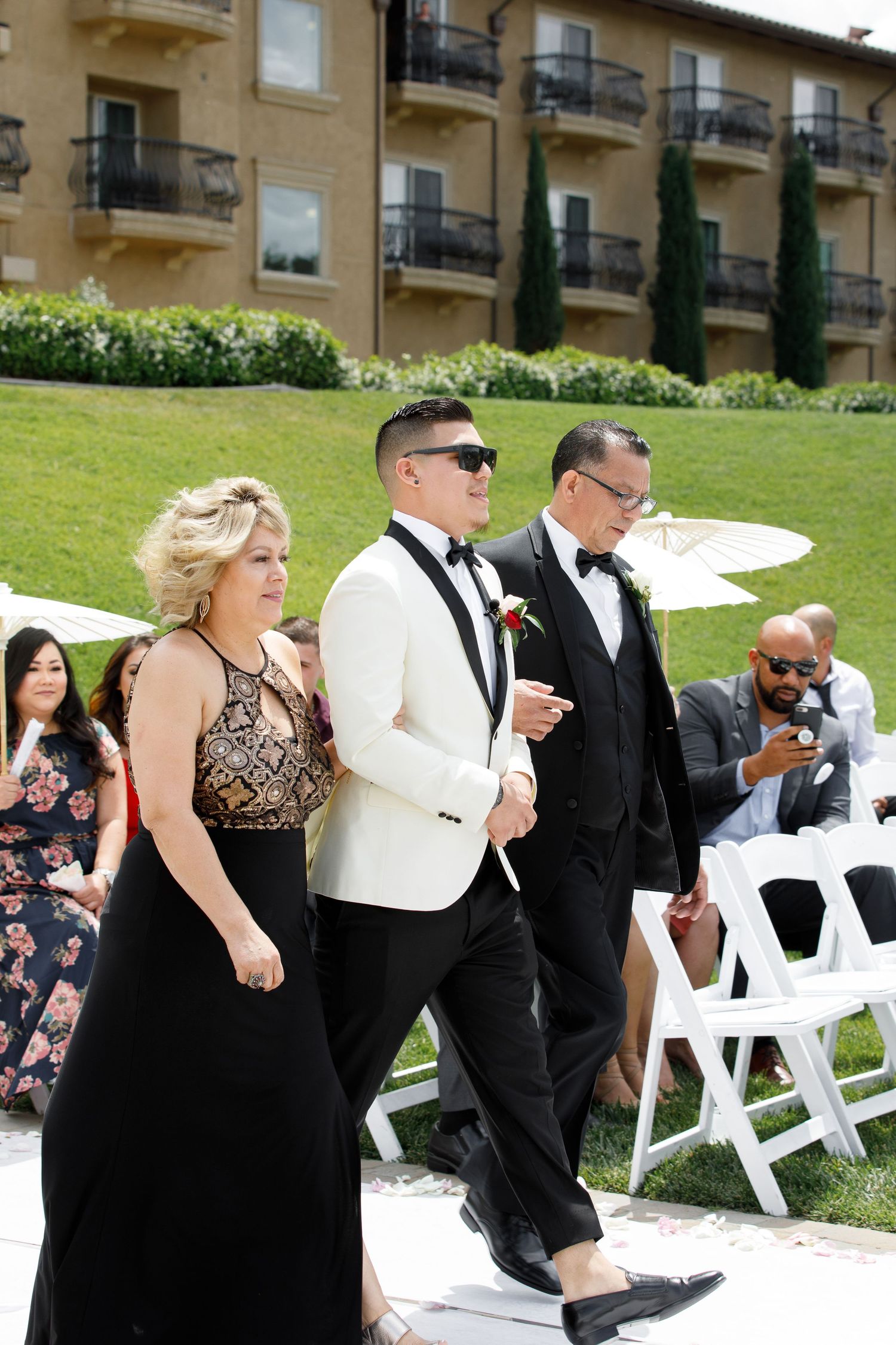 Wedding party members in formal black and white attire walk together in a line on a sunny day outdoors.