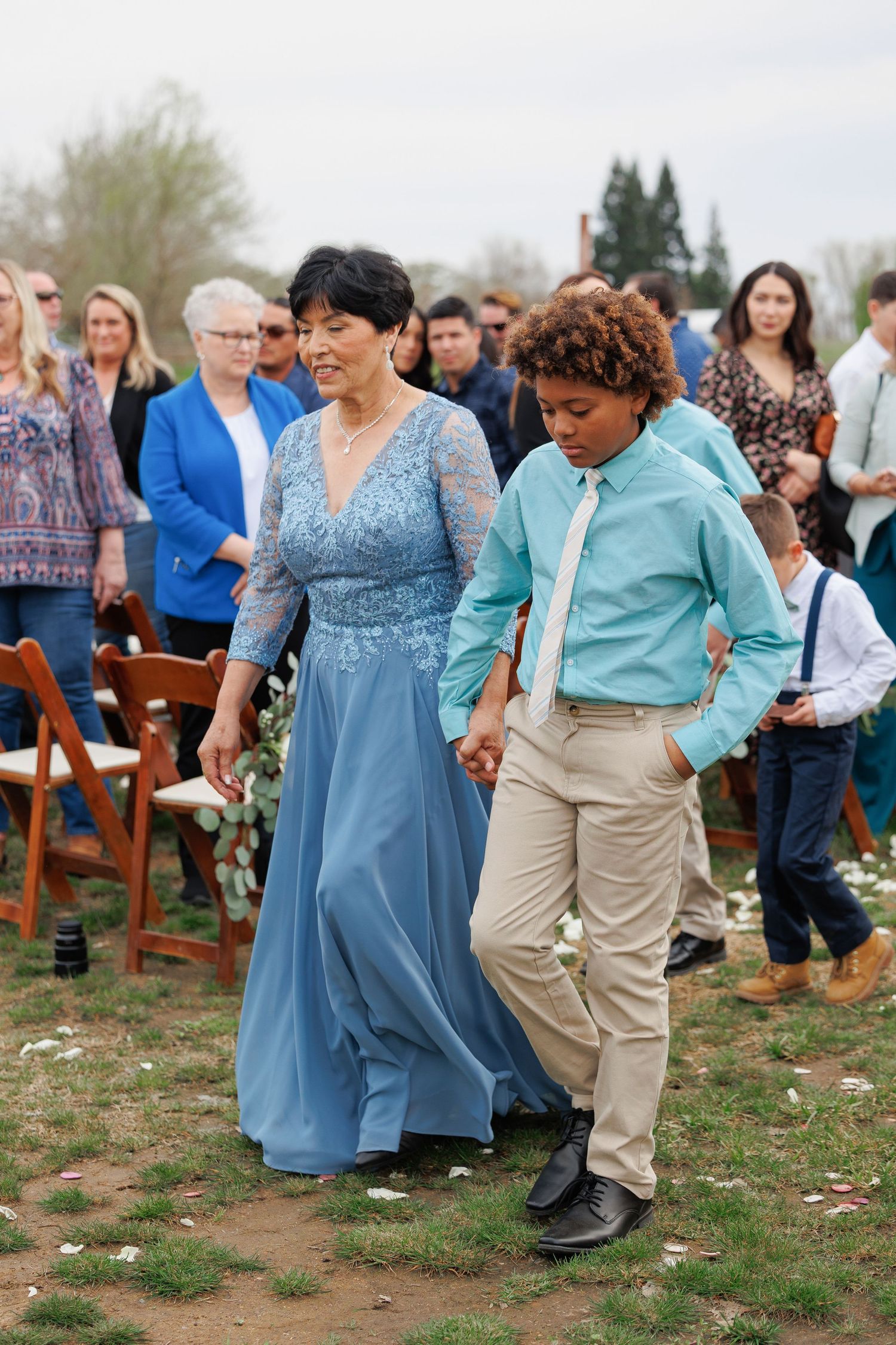 A group of wedding guests in blue dresses and khaki attire dance together at an outdoor celebration.